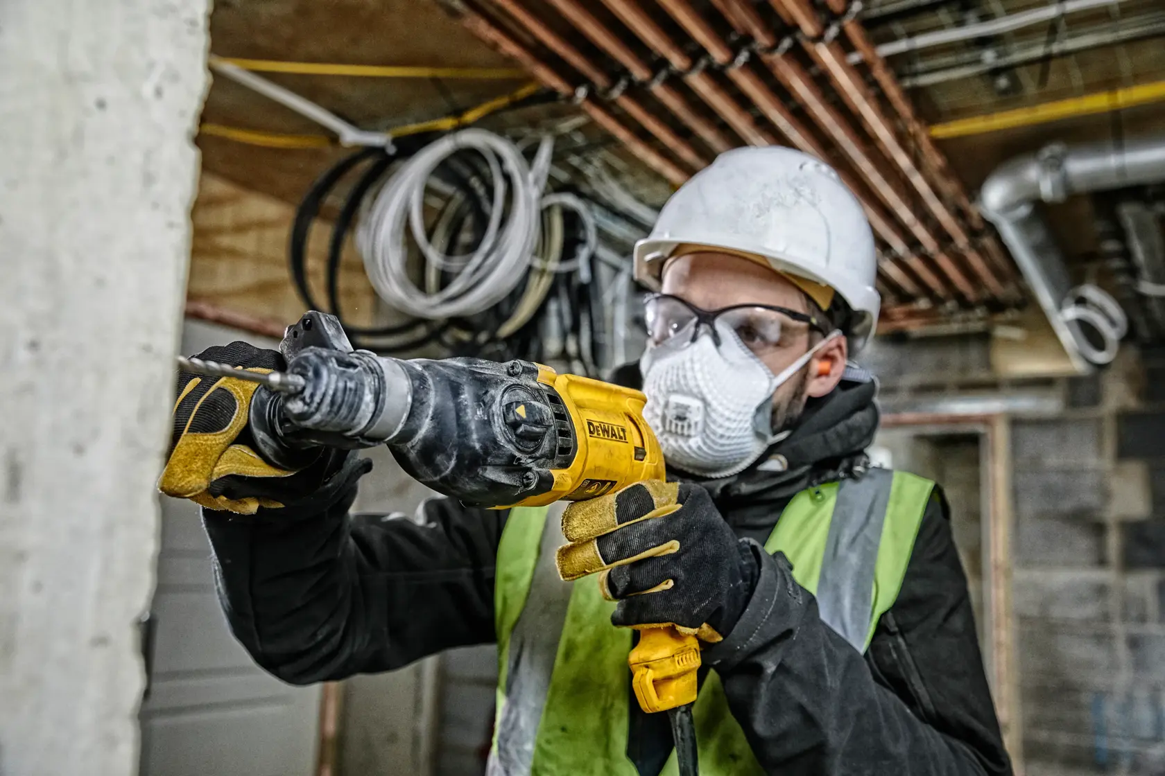 Ein Bauarbeiter mit Schutzhelm, Schutzbrille, Handschuhen und Warnweste benutzt einen DEWALT D25033 Bohrhammer auf einer Baustelle. Im Hintergrund sind unfertige Wände, Rohre und elektrische Kabel zu sehen.