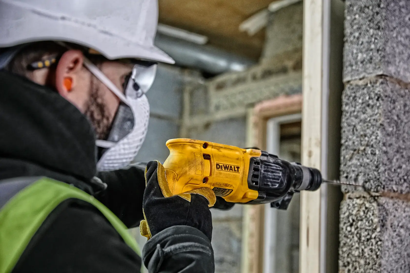 Eine Person mit Schutzausrüstung verwendet einen gelben DEWALT-Bohrhammer, um eine Betonwand auf einer Baustelle zu bohren.