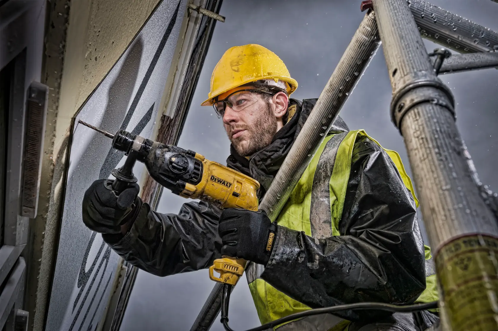 Ein Bauarbeiter mit gelbem Schutzhelm und Sicherheitsweste benutzt eine DEWALT Schlagbohrmaschine (Modell D25033), um eine Wand vom Gerüst aus zu bohren. Das Gesicht des Arbeiters ist aus Datenschutzgründen unscharf dargestellt.