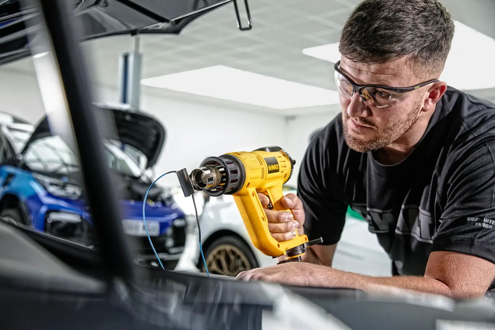 Eine Person verwendet eine gelbe DEWALT D26414 Heißluftpistole in einer Garage oder Werkstatt, im Hintergrund sind Autos zu sehen. Das Gesicht der Person ist zum Schutz der Privatsphäre verschwommen.