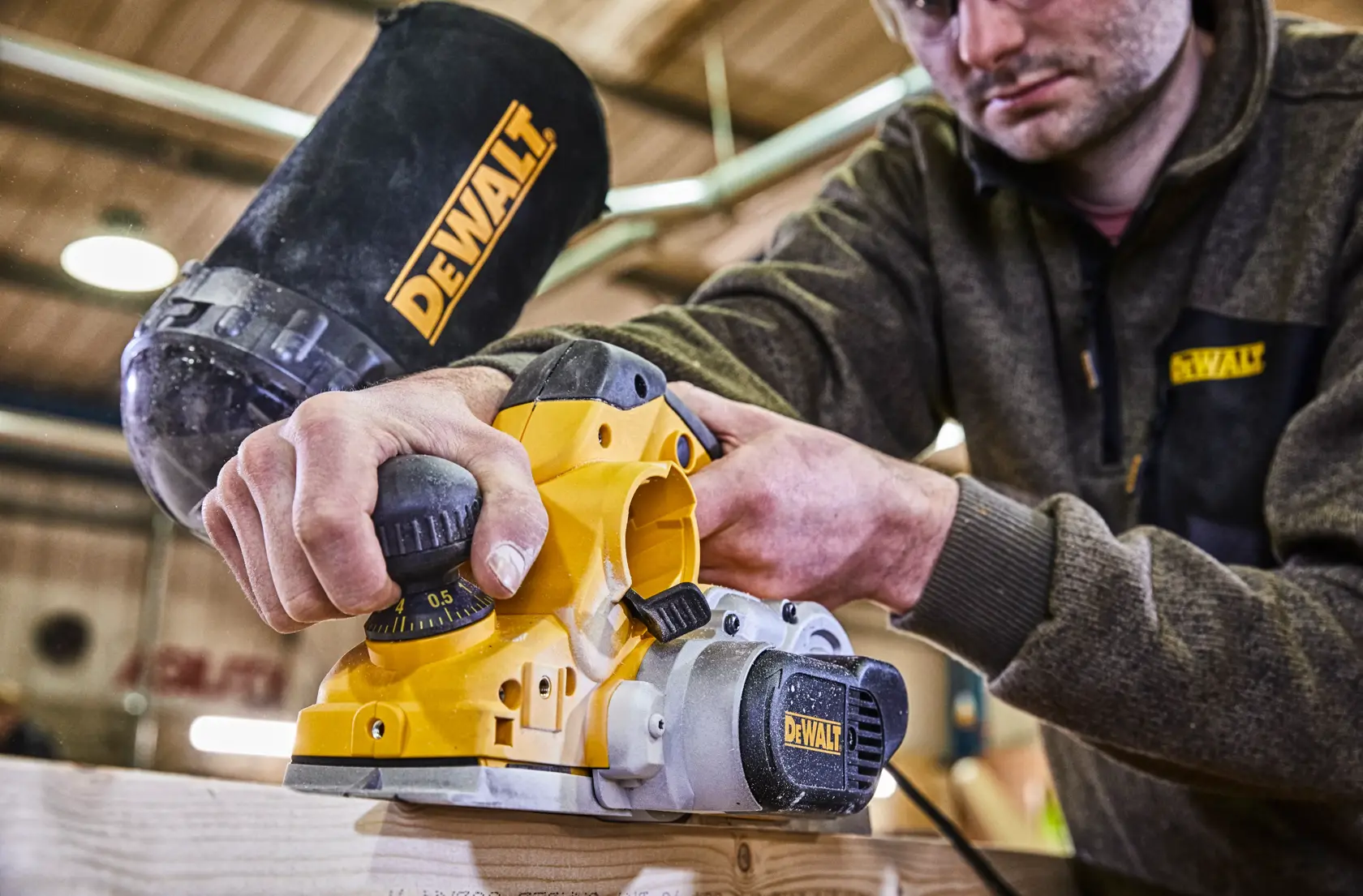 Eine Person verwendet einen elektrischen DEWALT Hobel, Modell D26500K, um ein Stück Holz zu glätten. Der Hobel ist gelb und schwarz mit sichtbarem DEWALT-Logo und die Szene spielt in einer Werkstatt. Das Gesicht der Person ist aus Datenschutzgründen verschwommen.