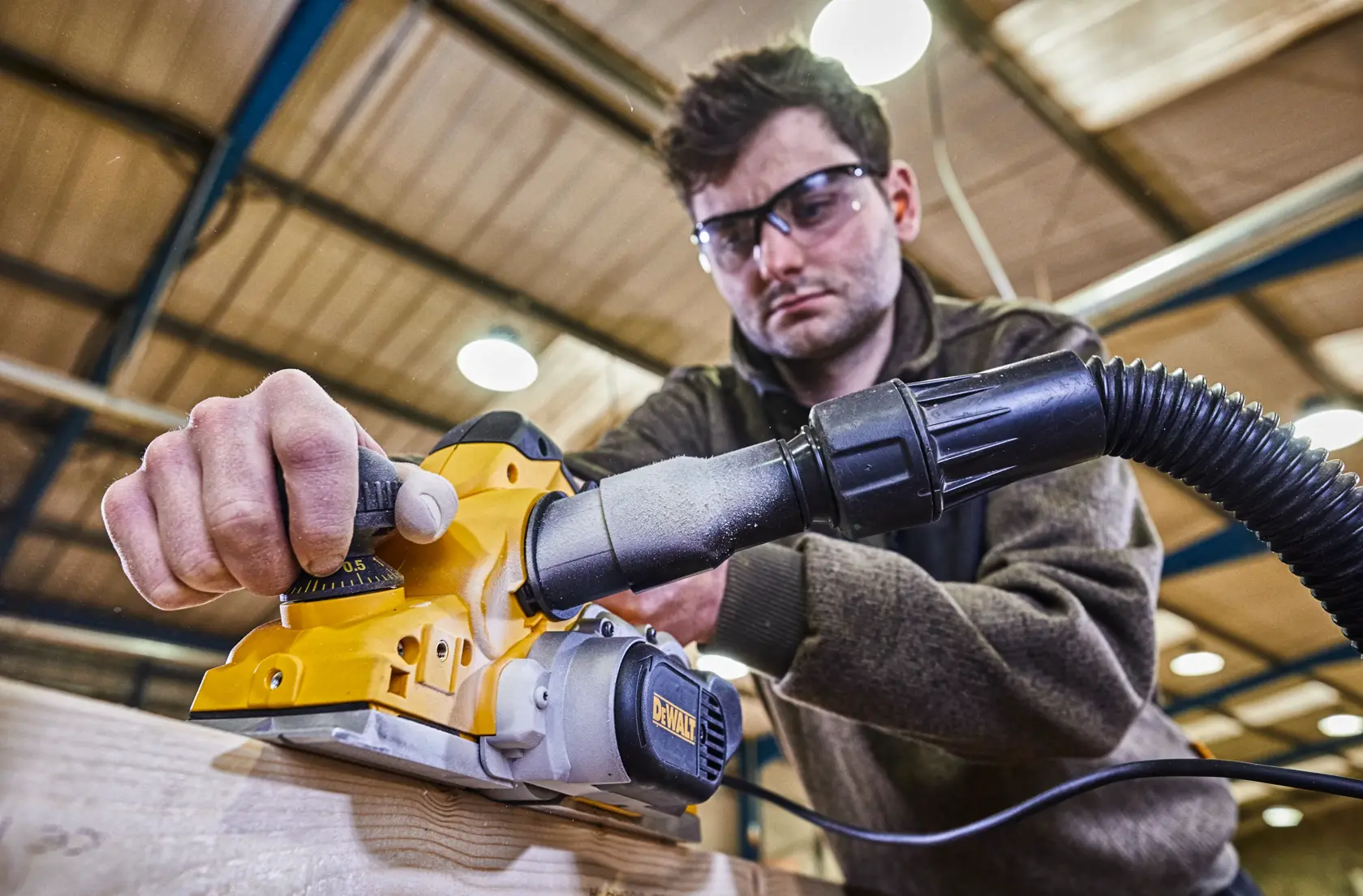 Una persona utiliza una cepilladora eléctrica DEWALT D26500 sobre un trozo de madera en un taller interior. El rostro de la persona está difuminado y la cepilladora está conectada a una manguera para extracción de polvo.