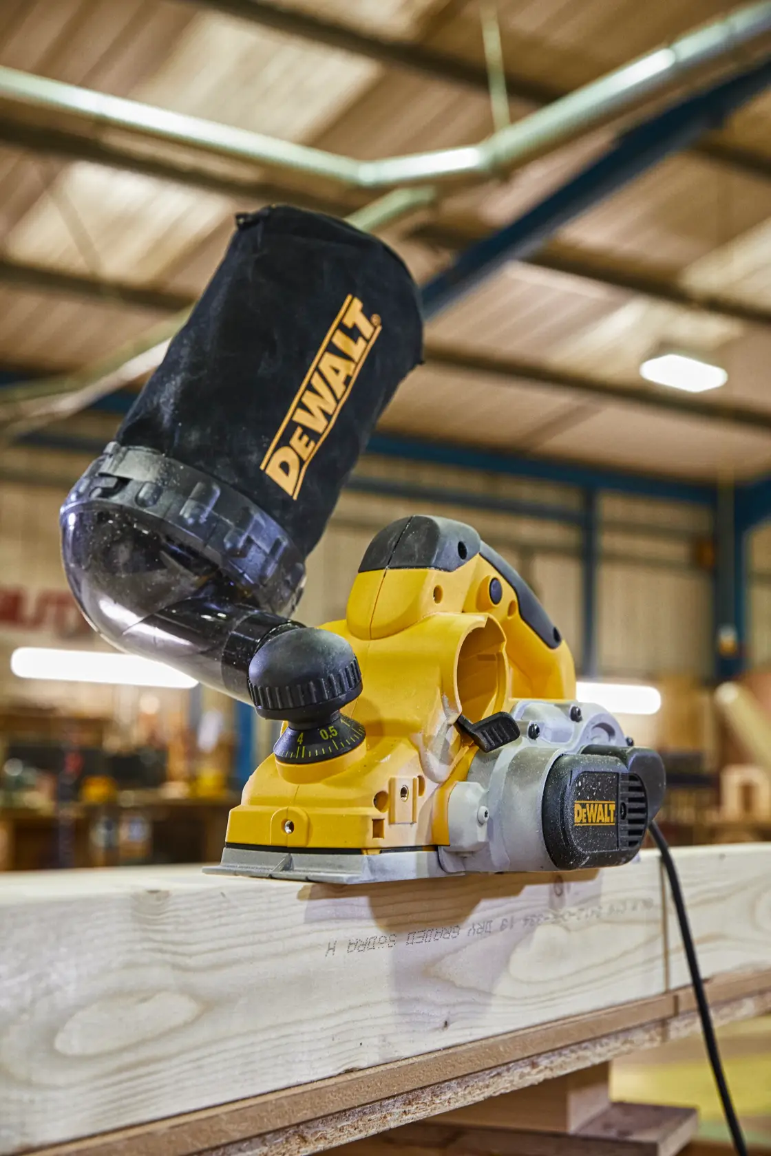 Se muestra una cepilladora eléctrica DEWALT en uso, alisando una tabla de madera en un taller. La cepilladora tiene una bolsa para el polvo en la parte superior para recoger las virutas de madera.