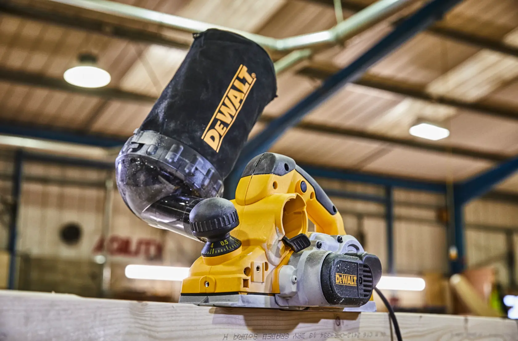 Una cepilladora eléctrica amarilla DEWALT con bolsa para el polvo adjunta, colocada sobre una superficie de madera dentro de un taller.
