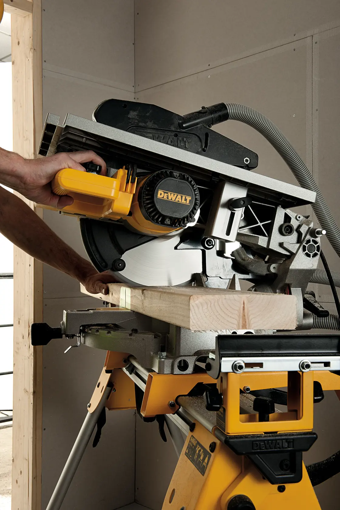 Eine Person verwendet eine DEWALT D27113 Säge, um ein Holzstück zu schneiden. Die Säge ist auf einem gelben Ständer montiert und wird in einer Werkstatt benutzt.
