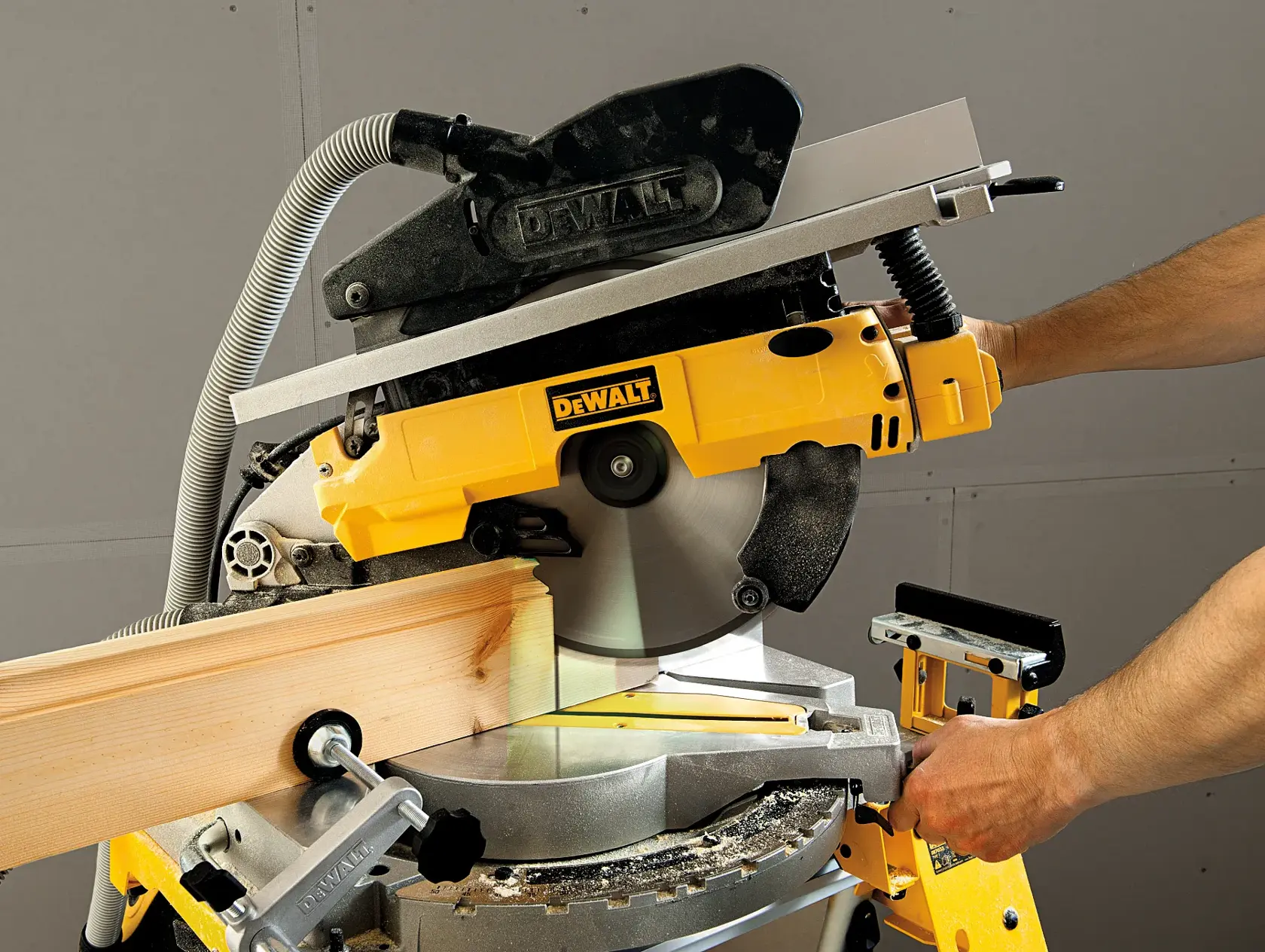 Eine Person benutzt eine DEWALT-Kapp- und Gehrungssäge (Modell D27113), um ein Stück Holz zu schneiden. Die Säge ist gelb und auf einer Werkbank montiert.