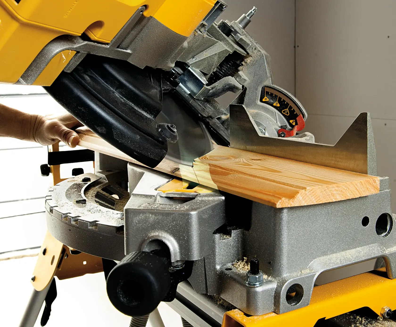 Nahaufnahme einer DEWALT Gehrungssäge, die ein Holzstück im Winkel schneidet. Die Säge ist auf einer Werkbank montiert und eine Hand hält das Holzstück fest. Auf der Oberfläche ist Sägemehl sichtbar.