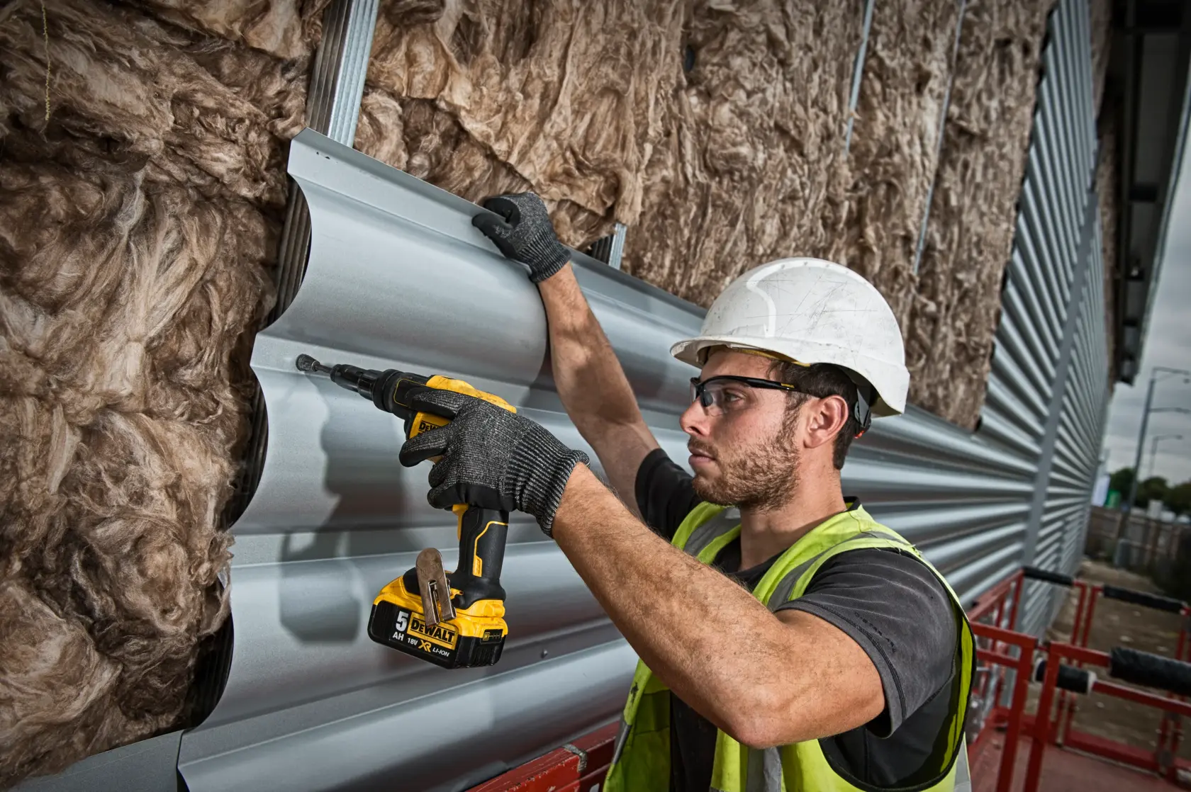 A construction worker wearing a white hard hat and gloves uses a DEWALT DCF622N cordless screwdriver to fasten metal panels onto an insulated wall at an industrial site.