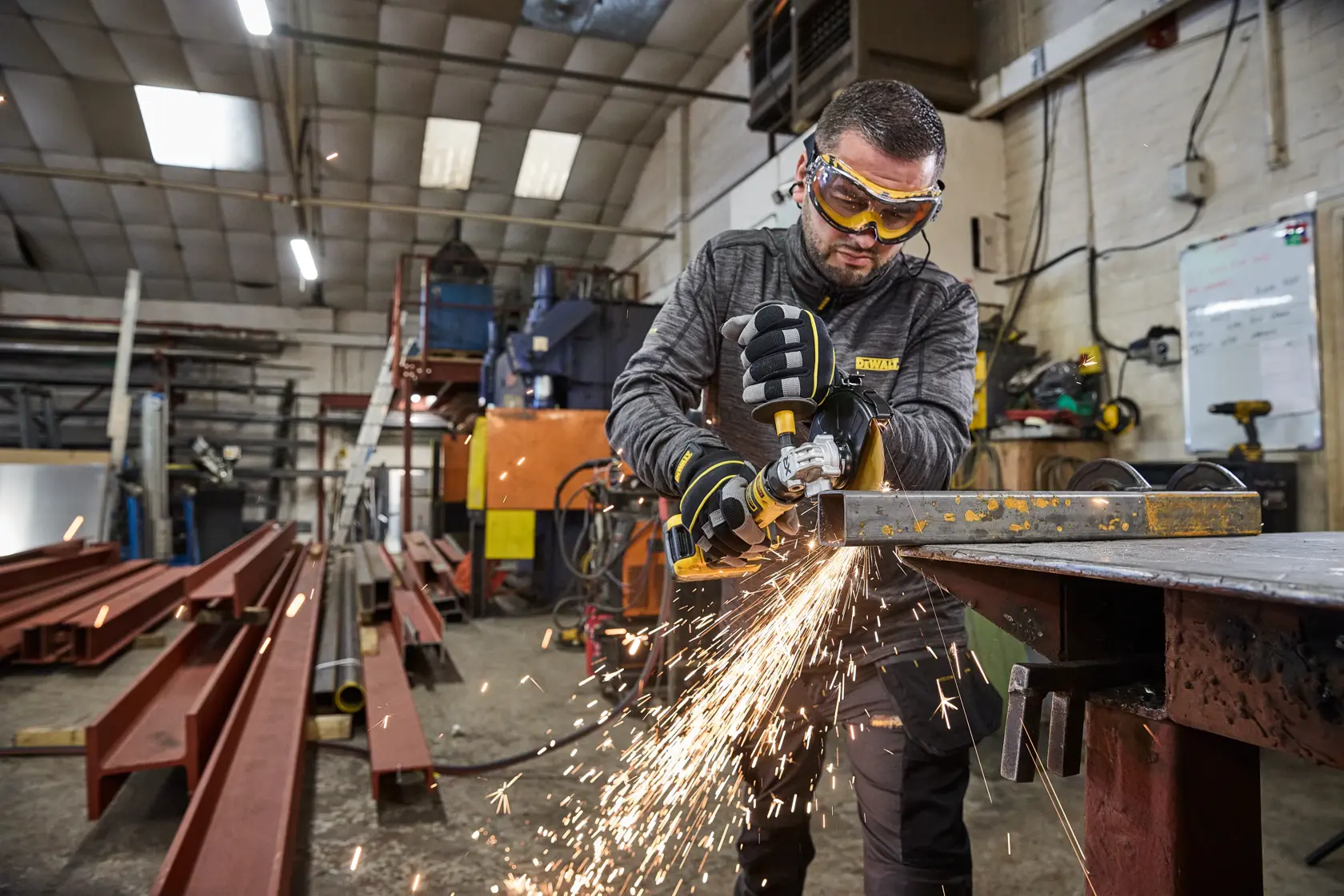 Person arbeitet mit DEWALT Winkelschleifer an Metall im Werkstatt; Funken fliegen. Industrieausrüstung vorhanden.