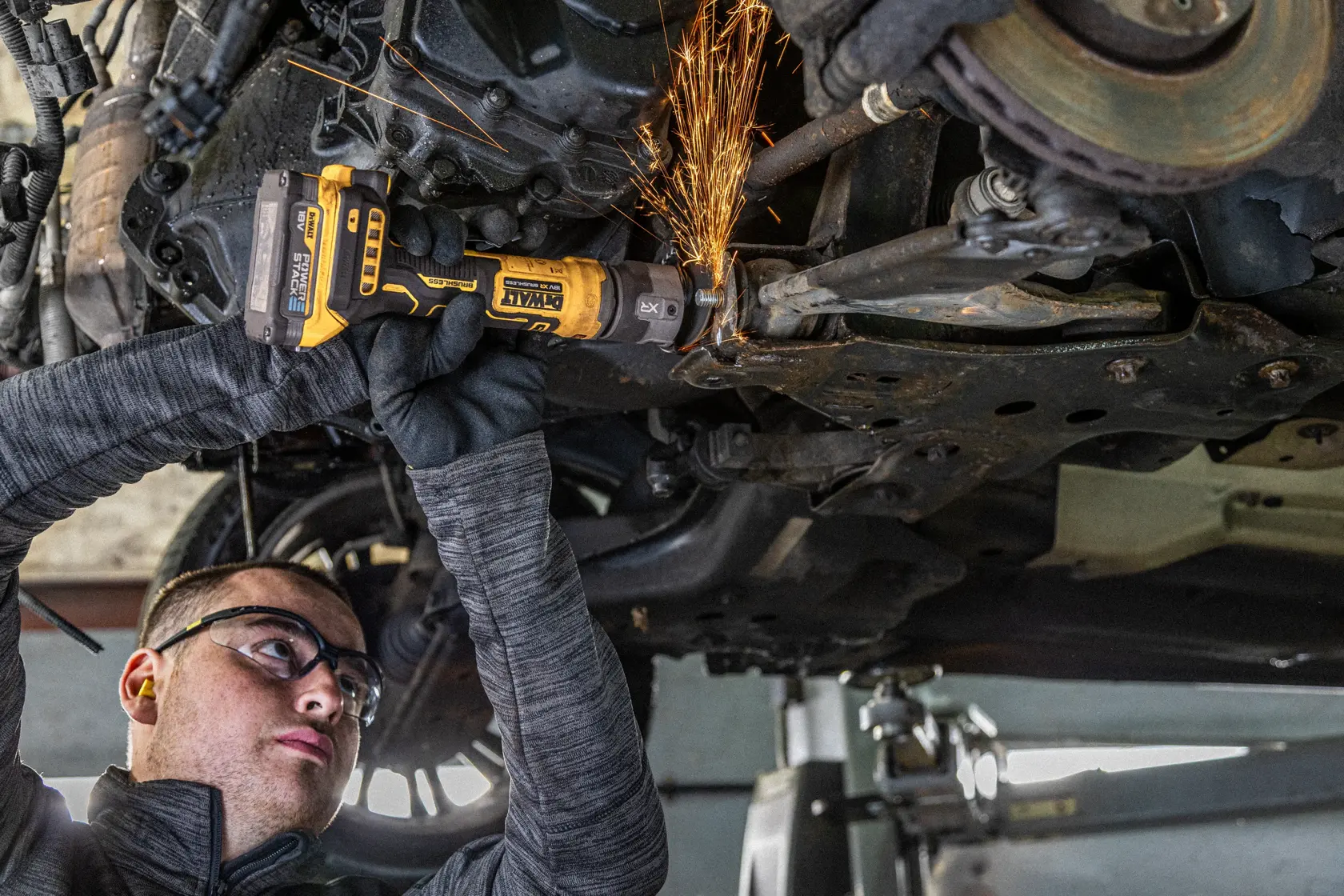 DEWALT DCG430N-XJ, DCG430N Schleifer unter Auto im Einsatz, Funken sichtbar, Werkzeug mit Handschuhen gehalten.