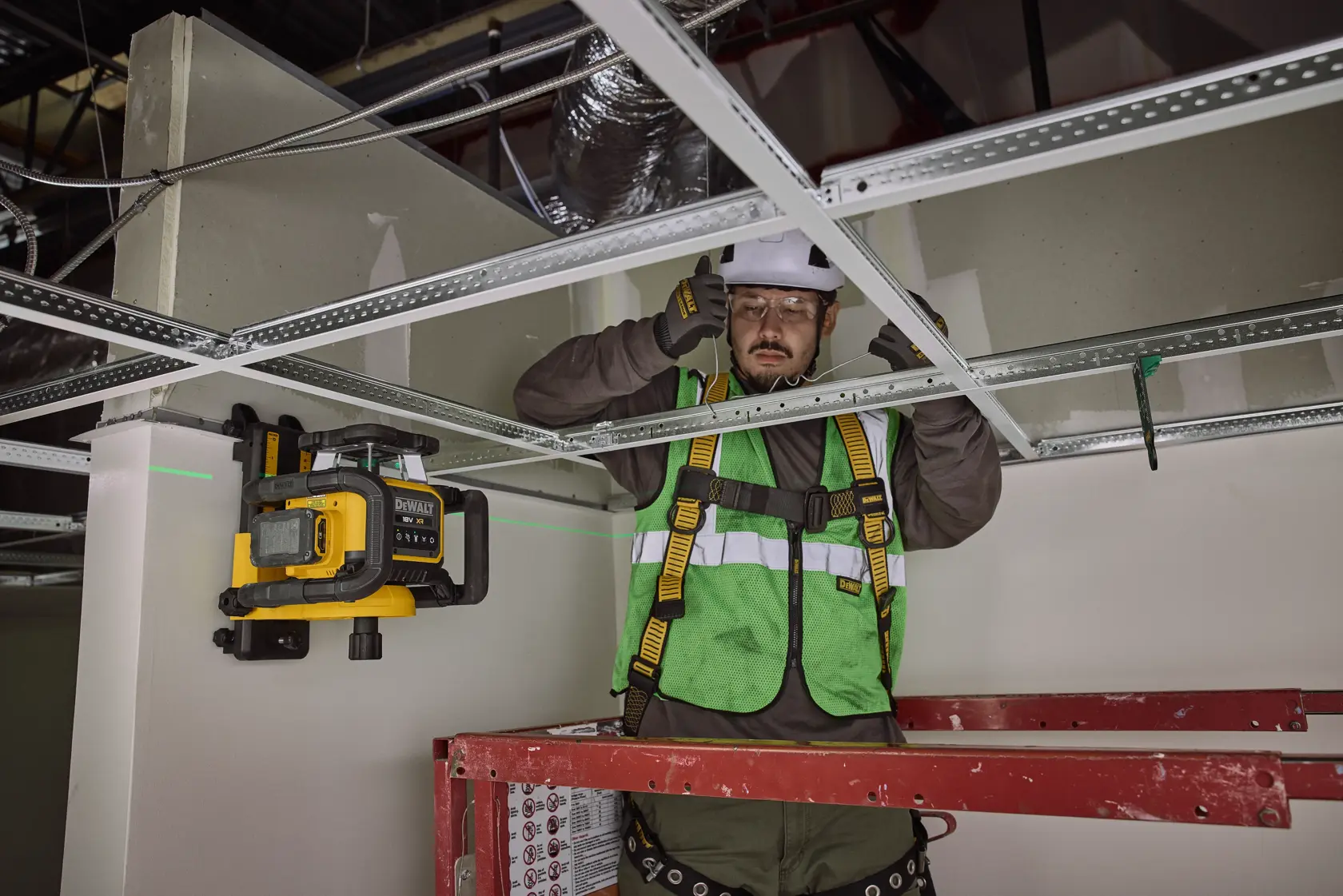 Arbeiter nutzt DEWALT Laserwerkzeug für Deckenmontage, trägt Schutzkleidung und steht auf einer Leiter.