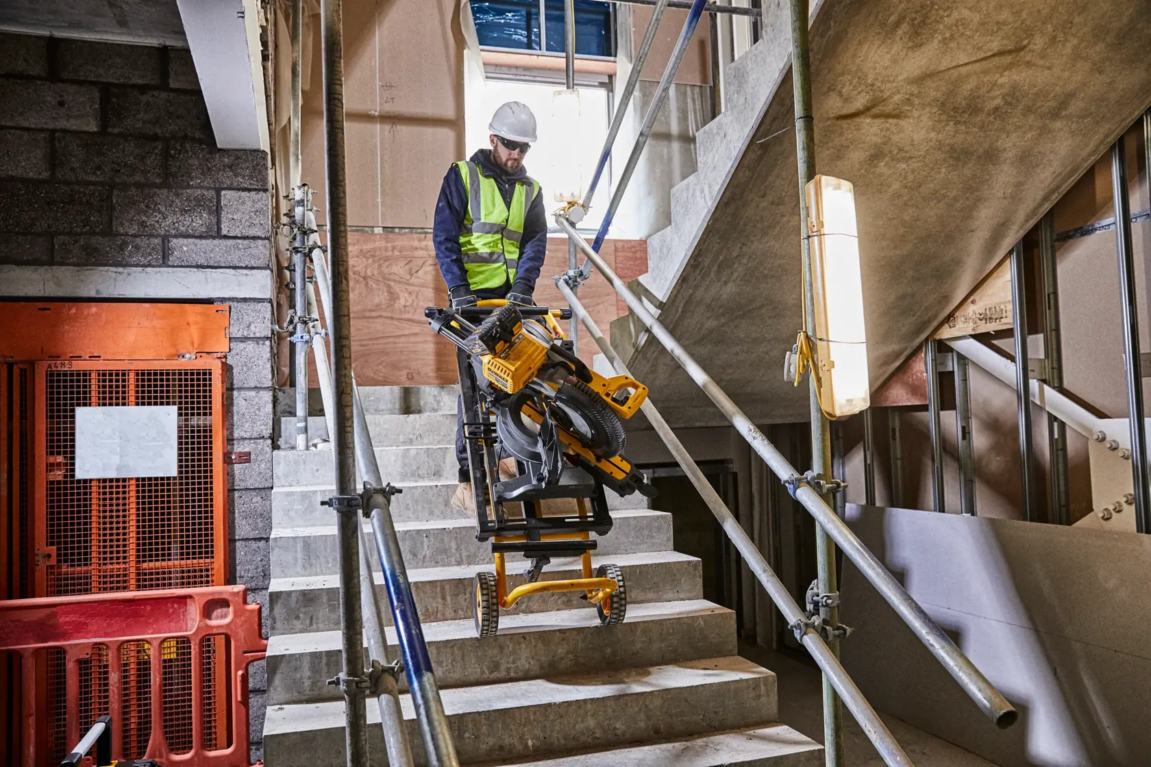 Arbeiter transportiert DEWALT Säge und Gestell auf Treppe einer Baustelle mit Sicherheitsgeländern.