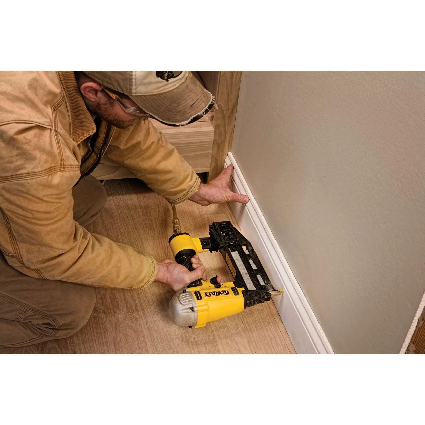 Person befestigt mit DEWALT Nagelgerät eine Leiste an einer Wand in einem Raum mit Holzfußboden.