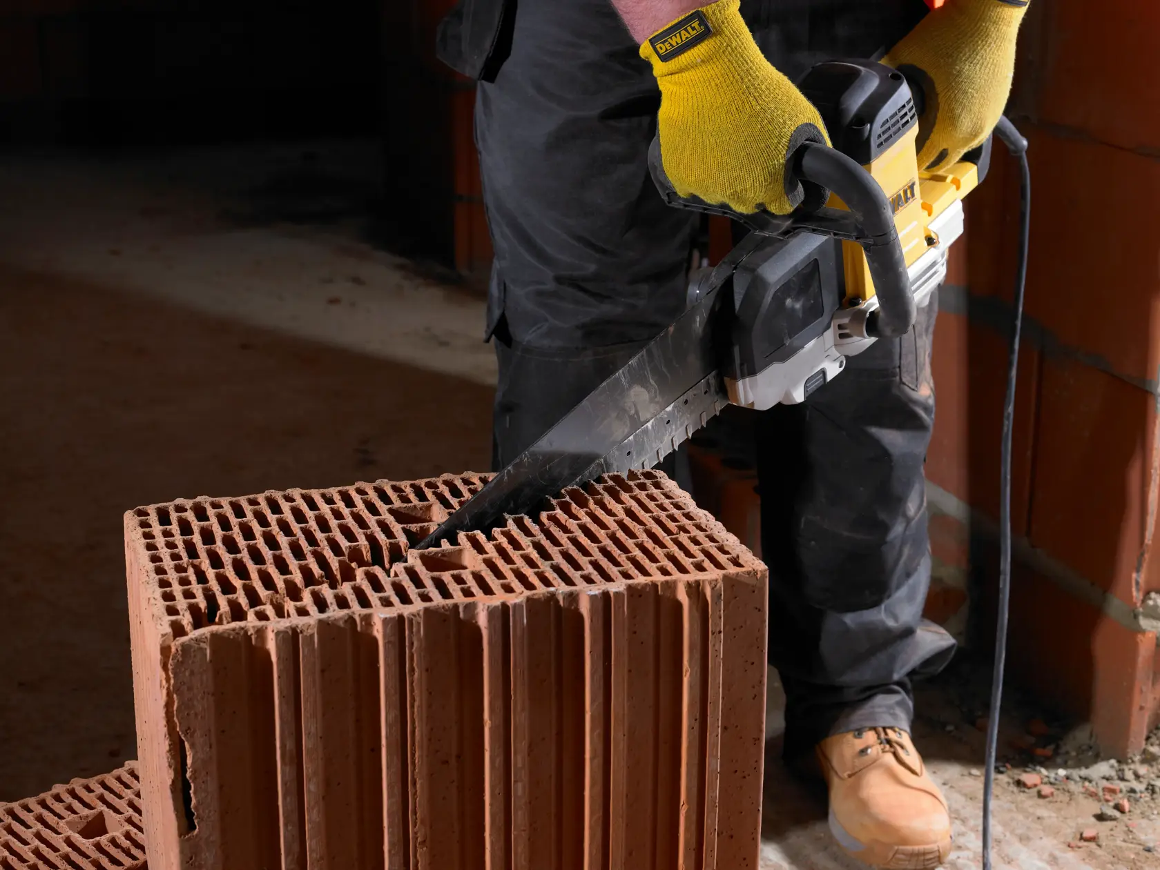 Persona corta un bloque de ladrillo hueco con una sierra DEWALT. Guantes amarillos y botas visibles.