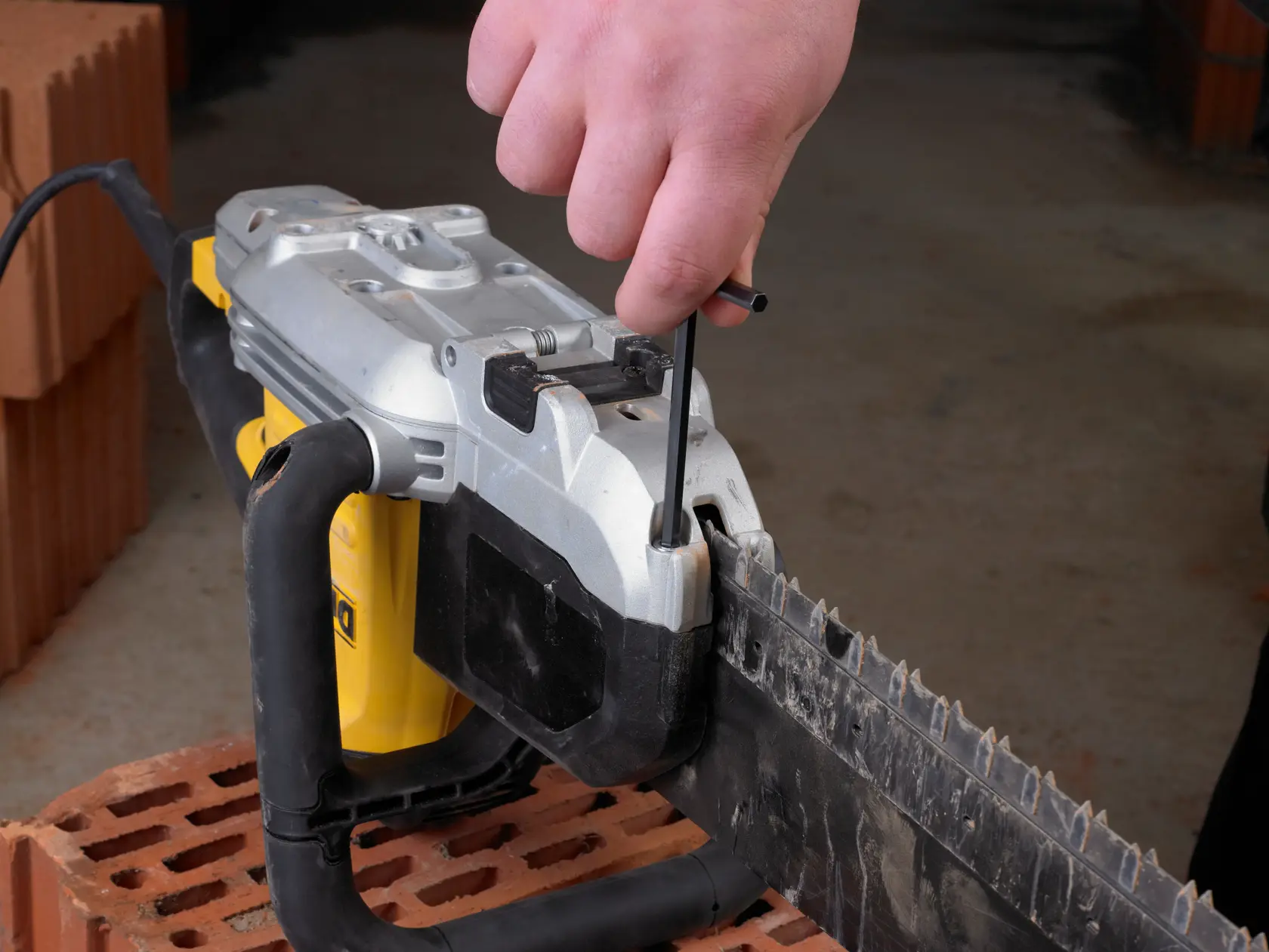 Mano ajusta el filo de una motosierra DEWALT usando herramienta sobre ladrillos, ambiente interior.