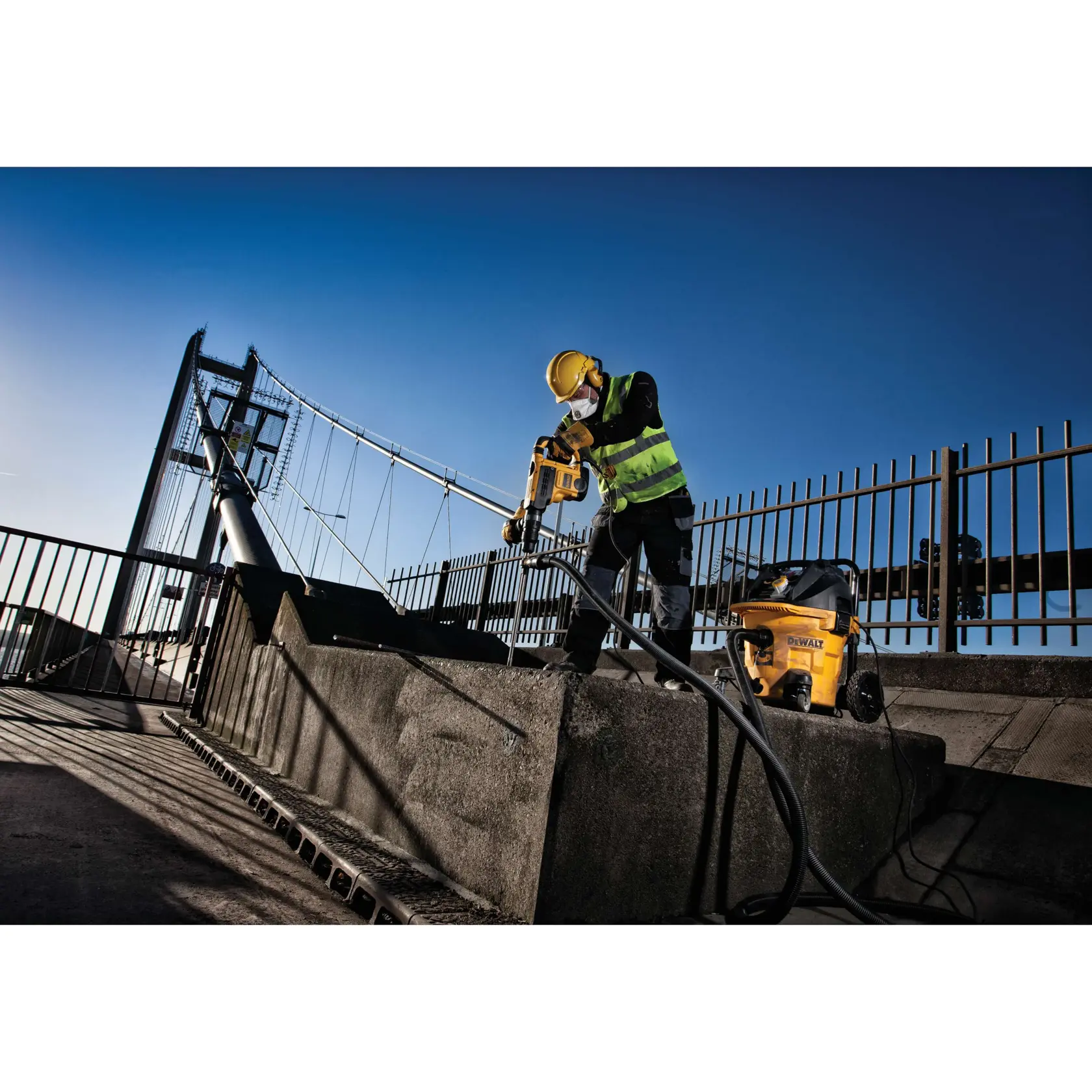 Trabajador usa taladro DEWALT en concreto al aire libre junto a un puente, con equipo de seguridad y chaleco.