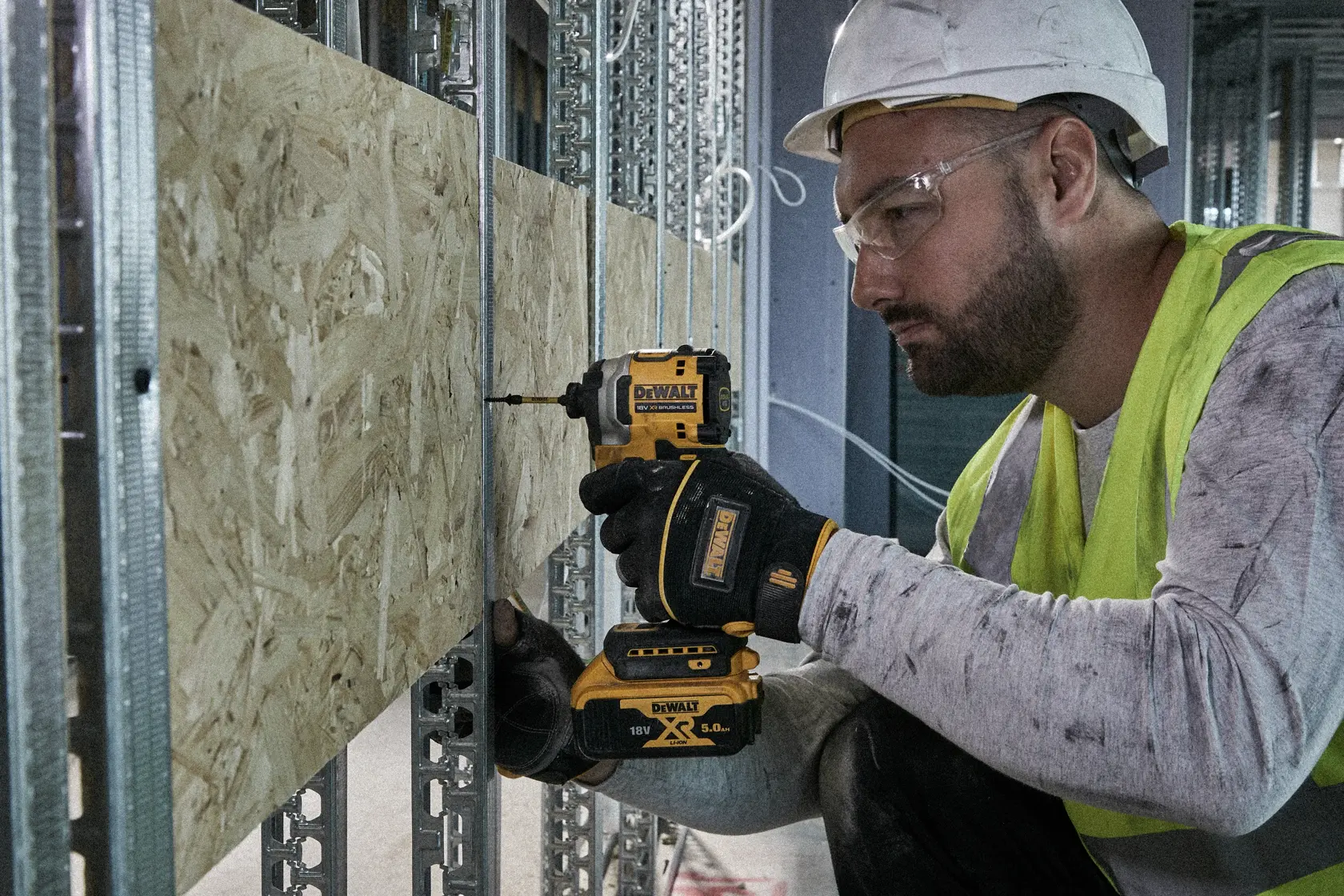 Person nutzt DEWALT-Bohrmaschine an Holzplatte auf Baustelle, mit Schutzkleidung und Handschuhen.