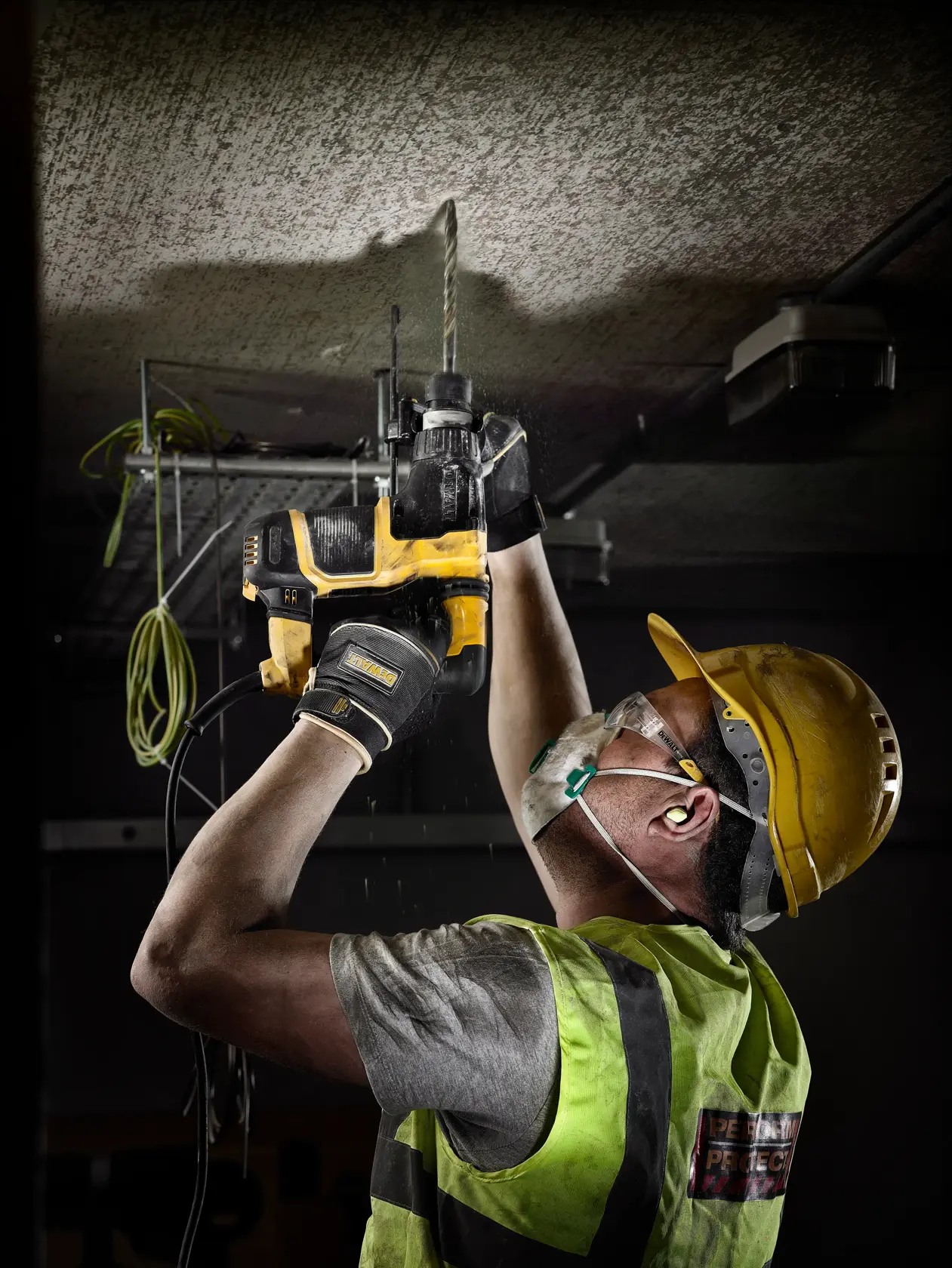 Arbeiter mit Schutzausrüstung bohrt mit DEWALT-Bohrmaschine in eine Decke auf der Baustelle.