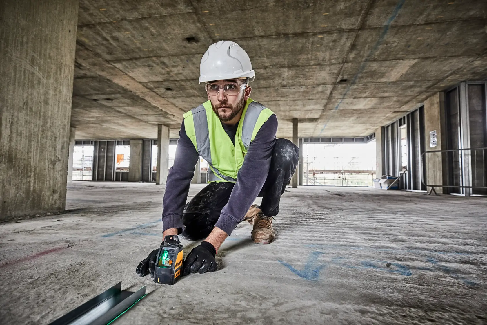 Eine Person mit Sicherheitshelm, Warnweste und Handschuhen kniet auf dem Boden einer großen Baustelle und verwendet einen