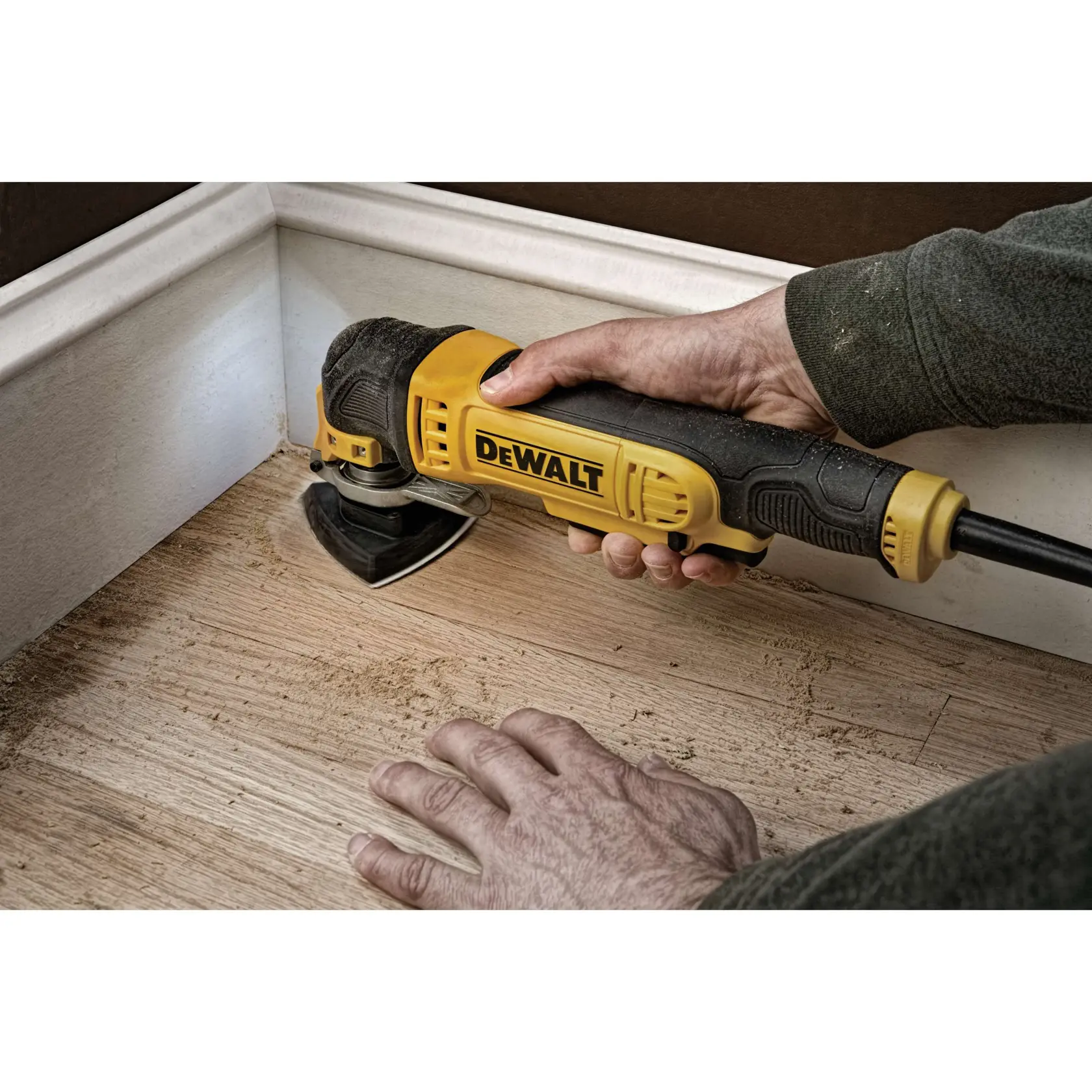Eine Person verwendet ein oszillierendes Multifunktionswerkzeug von DEWALT, um einen Holzboden in der Nähe einer Fußleiste zu schleifen oder zu schneiden.