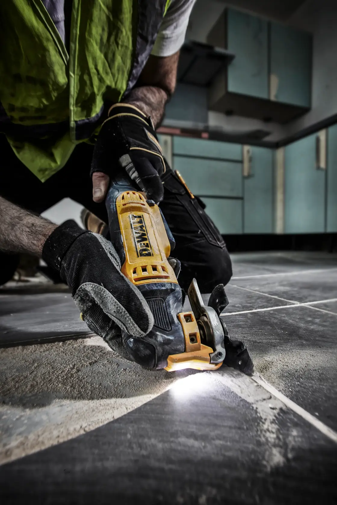 Hand mit Handschuhen nutzt DEWALT DWE315 Multitool zum Schneiden von Fliesenfugen im Küchenboden.