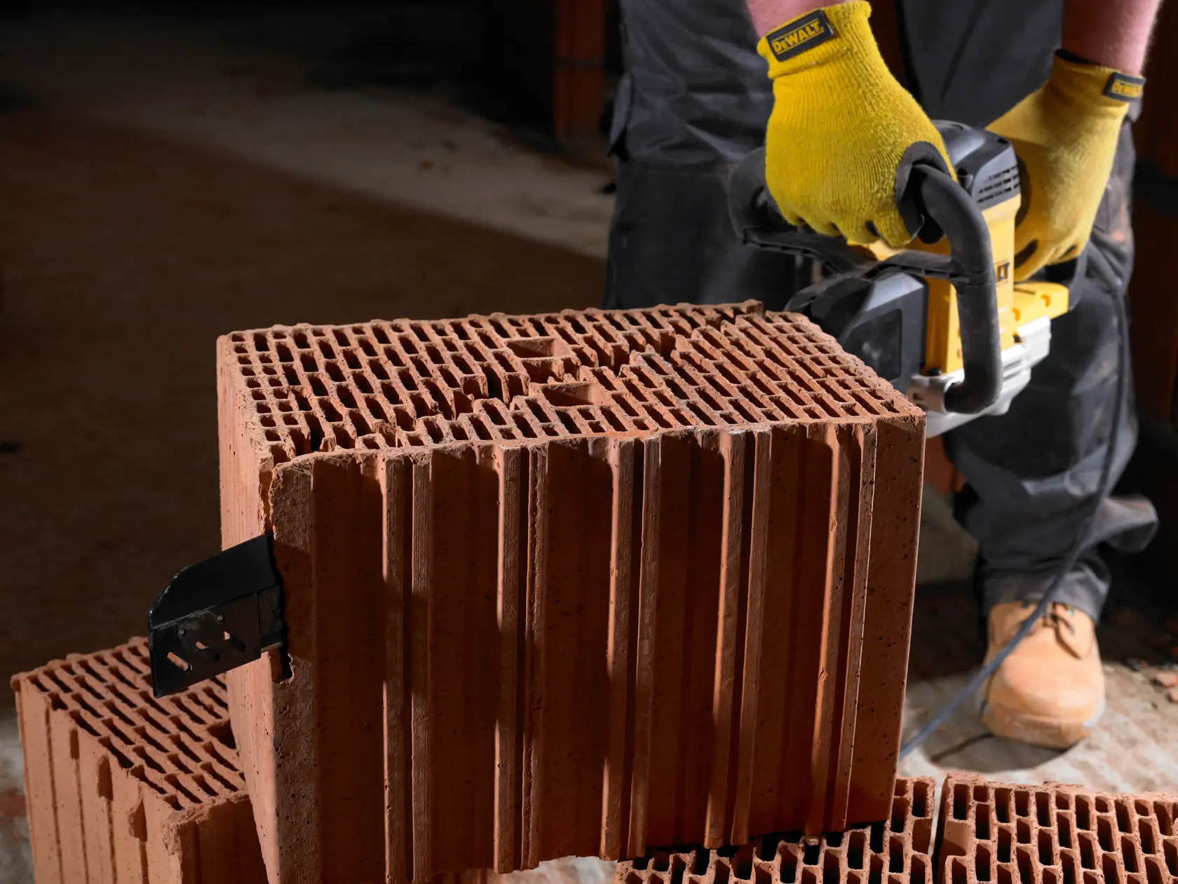 Eine Person mit gelben Handschuhen verwendet ein DEWALT Elektrowerkzeug, um einen großen, hohlen Tonziegel zu schneiden. Das Werkzeug ist gegen den Ziegel positioniert und die Person steht auf einer staubigen Oberfläche.