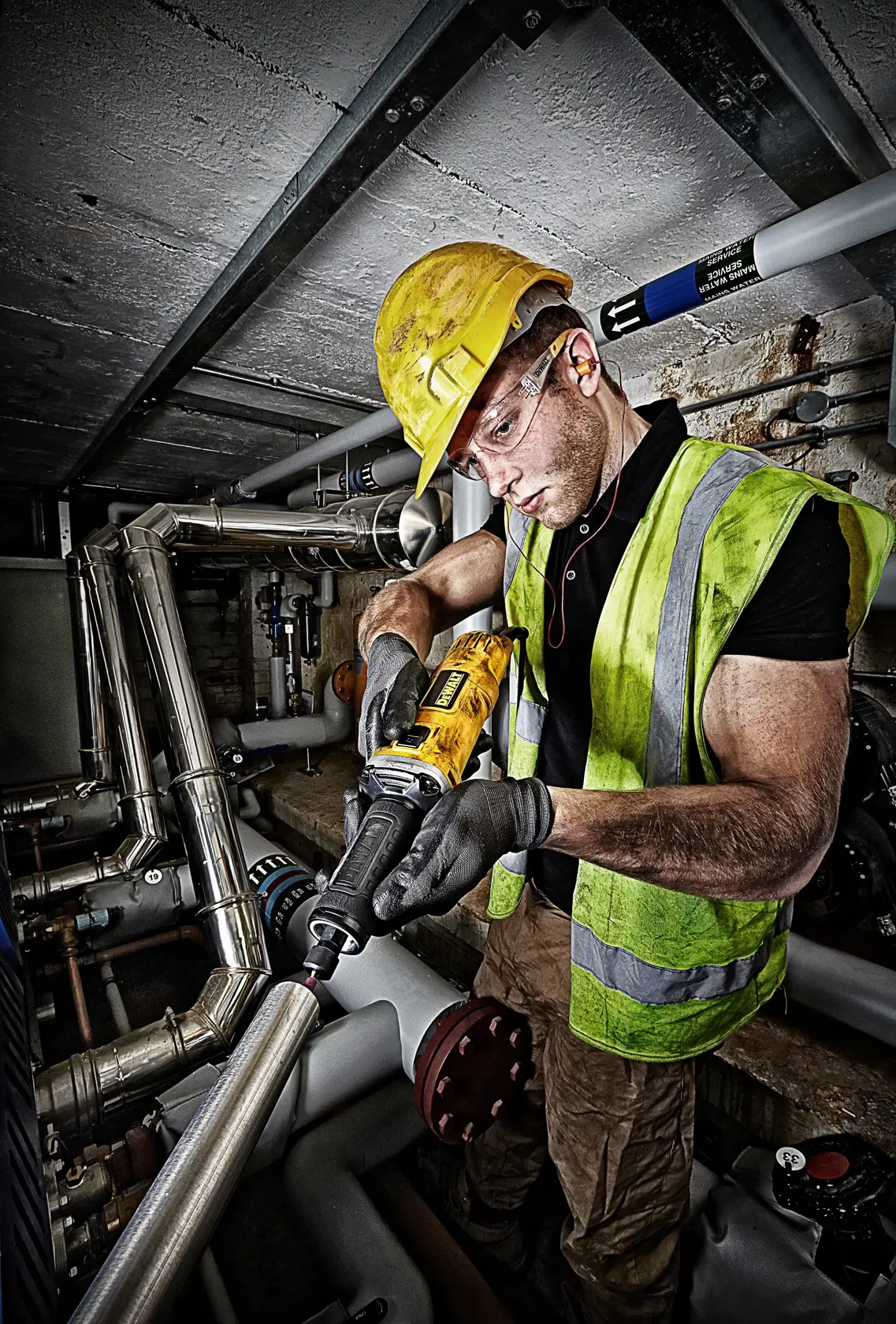 Arbeiter nutzt DEWALT DWE4884 Schleifer am Rohr und trägt Sicherheitskleidung in industrieller Umgebung.