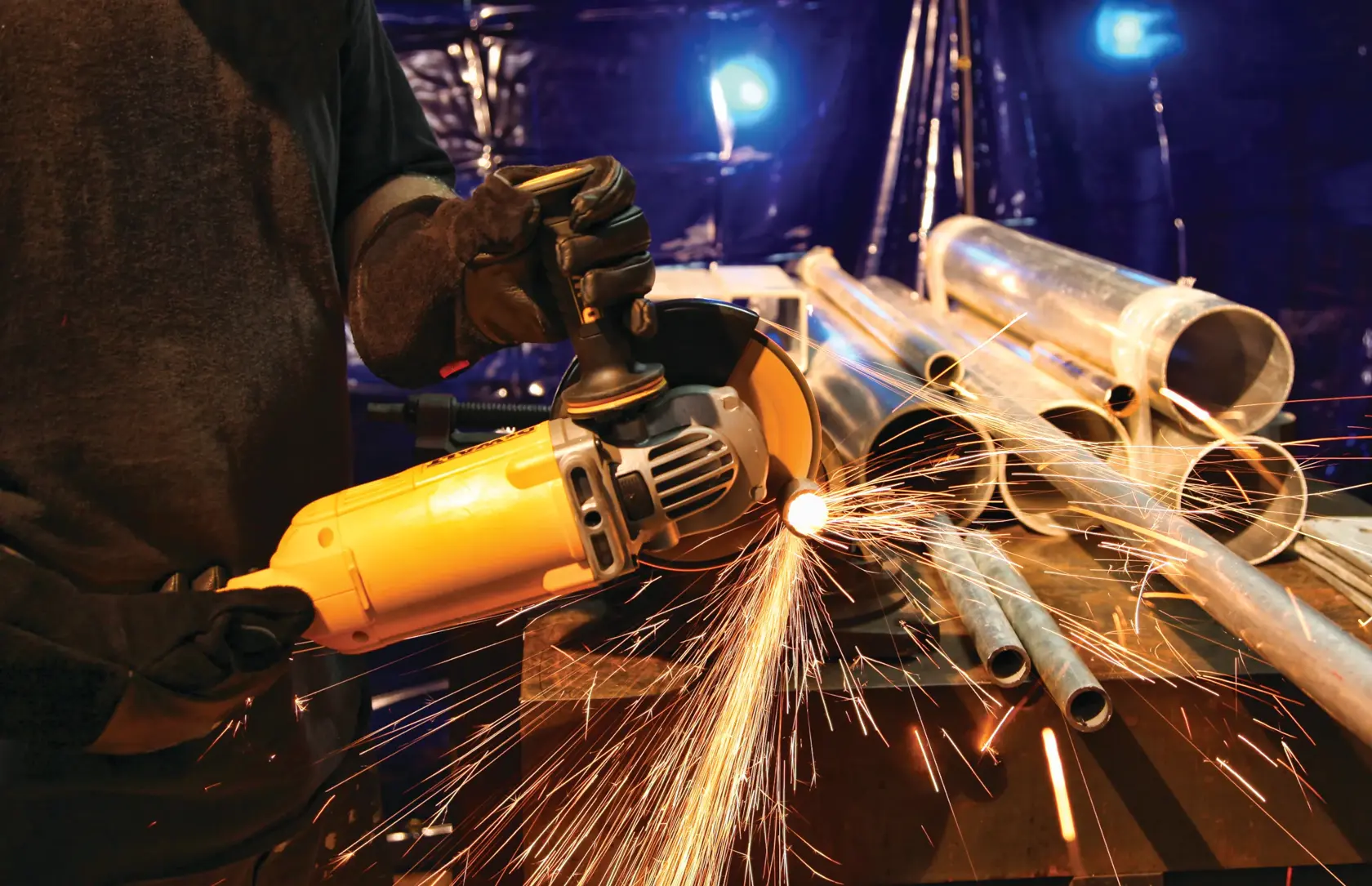 Eine Person mit Handschuhen benutzt einen gelben DEWALT-Winkelschleifer, um Metallrohre zu schneiden, während Funken vom Schneidebereich fliegen. Mehrere Metallrohre sind auf einem Arbeitstisch in einer Werkstatt gestapelt.