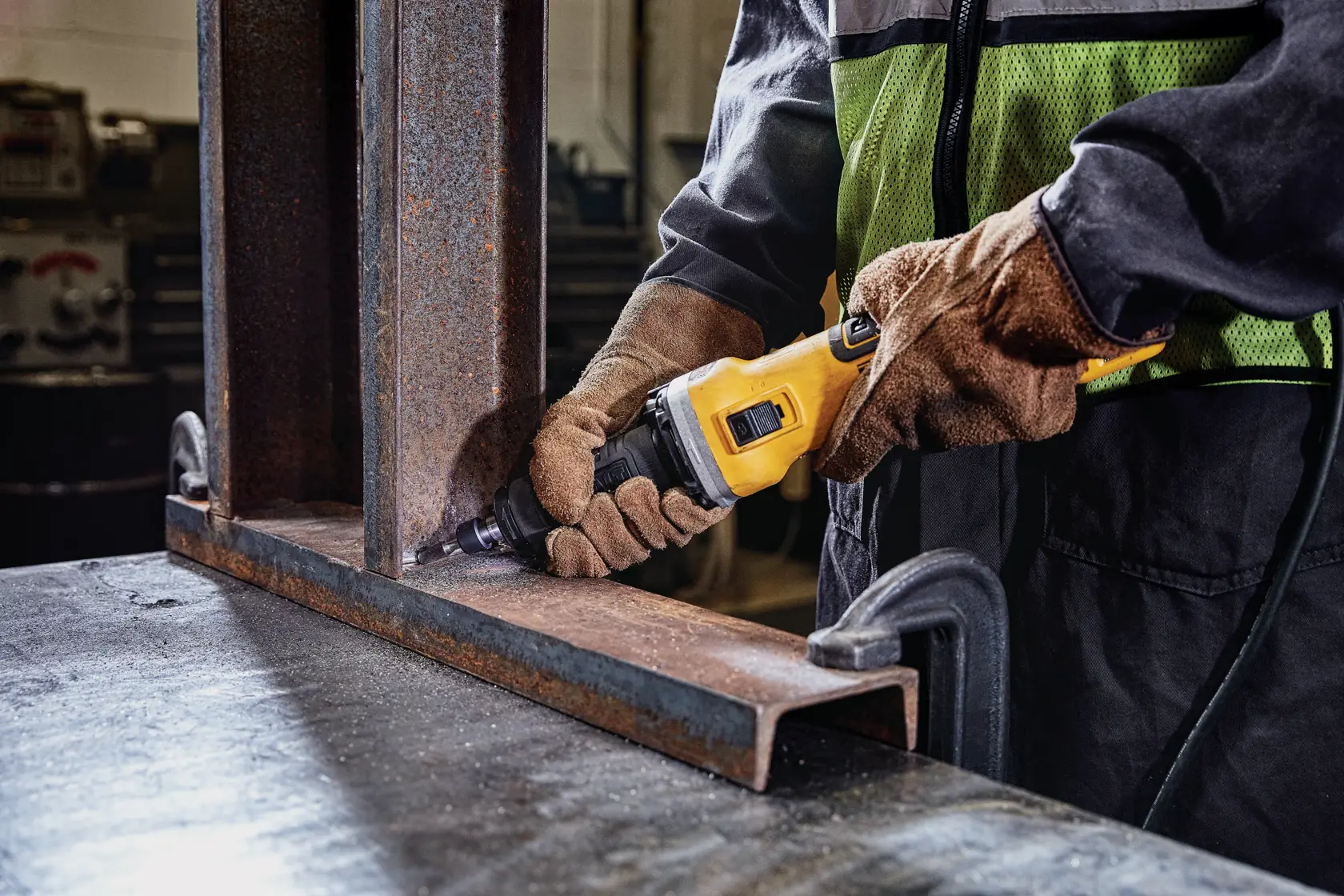 Person schleift Metallträger mit DEWALT Werkzeug, trägt Handschuhe und Schutzkleidung in Werkstatt.