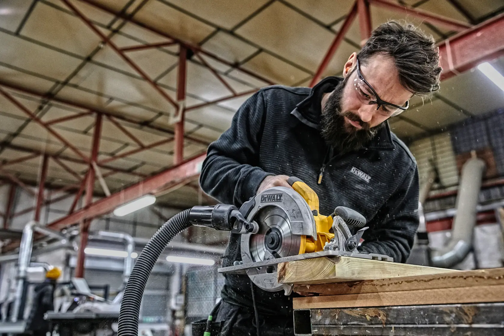 Arbeiter sägt Holz mit DEWALT DWE576K Kreissäge in Werkstatt, Staubsaugerschlauch angeschlossen.
