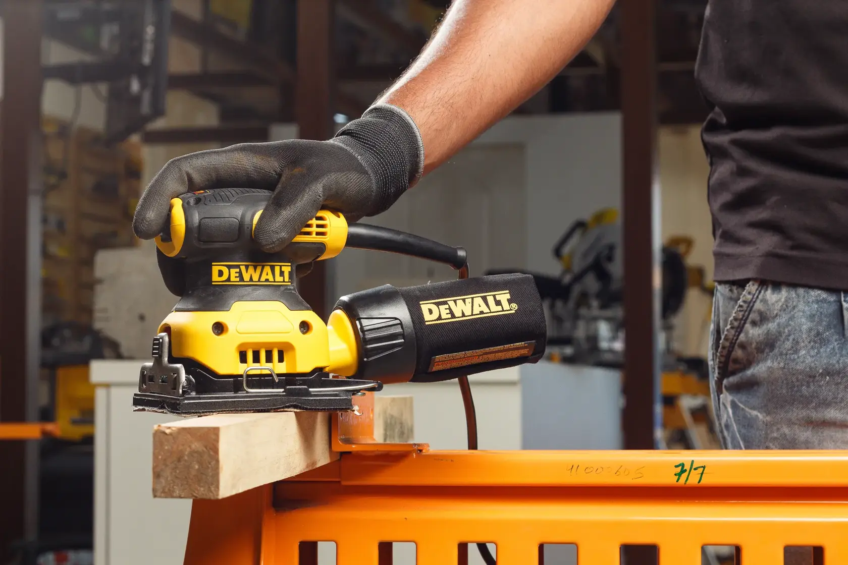 Nahaufnahme eines Menschen mit Arbeitshandschuhen, der einen gelben DEWALT-Handschleifer (SKU: DWE6411) verwendet, um die Oberfläche eines Holzbretts im Freien zu schleifen.