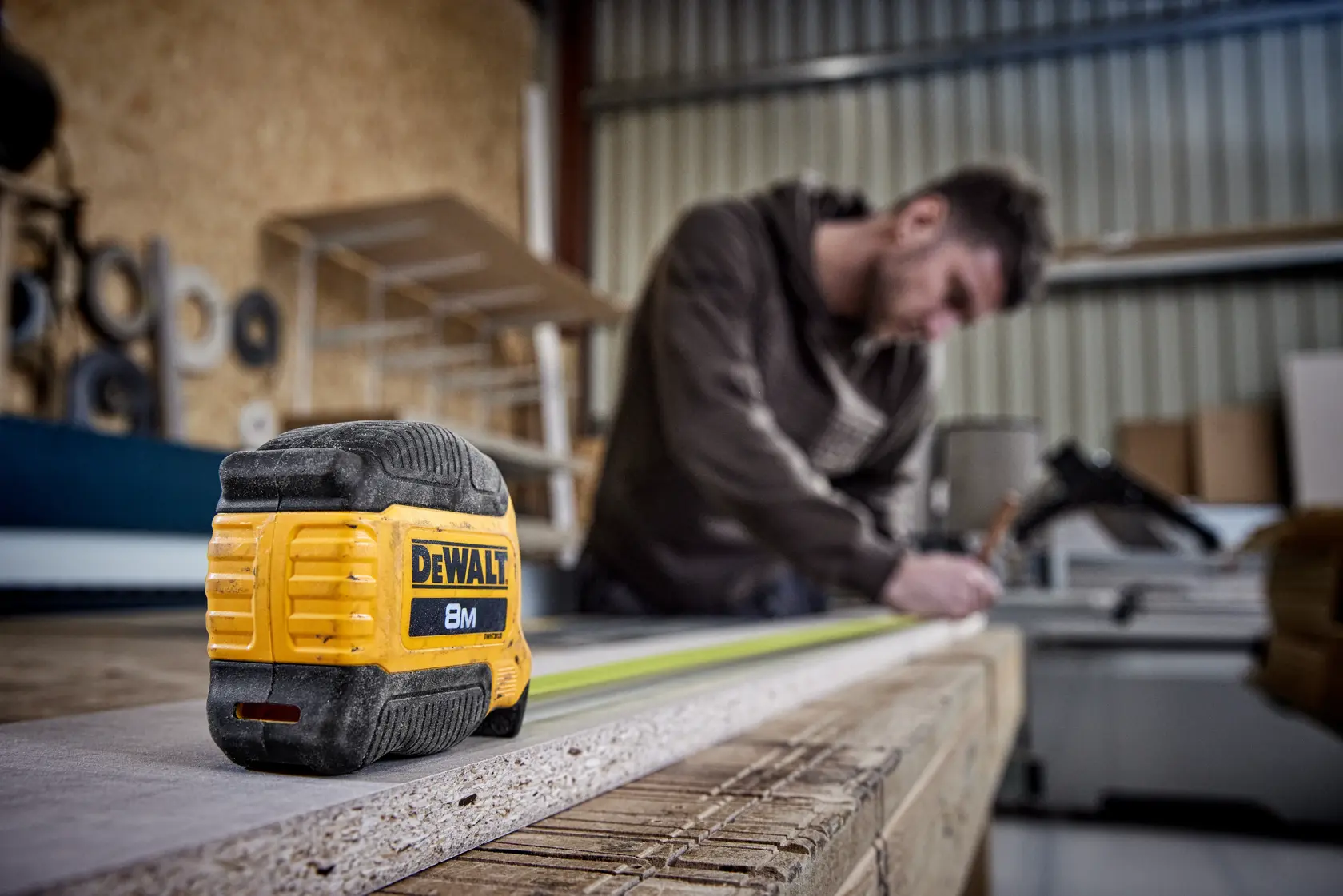 DEWALT 8M Maßband auf Holz, Person misst im Hintergrund in einer Werkstatt.
