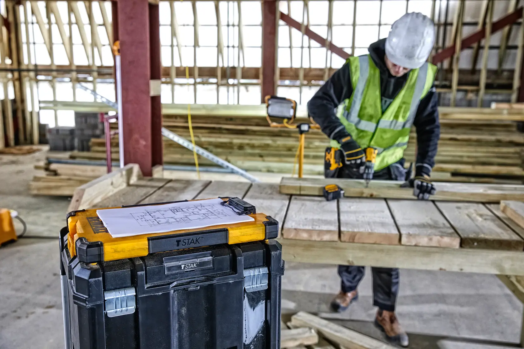 Ein DEWALT TSTAK-Werkzeugkasten im gelb-schwarzen Design steht im Vordergrund einer Baustelle. Auf dem Kasten liegt ein Blatt Papier mit Zeichnungen. Im Hintergrund arbeitet ein Bauarbeiter mit Schutzhelm und Warnweste mit einem Elektrowerkzeug an Holzbrettern.