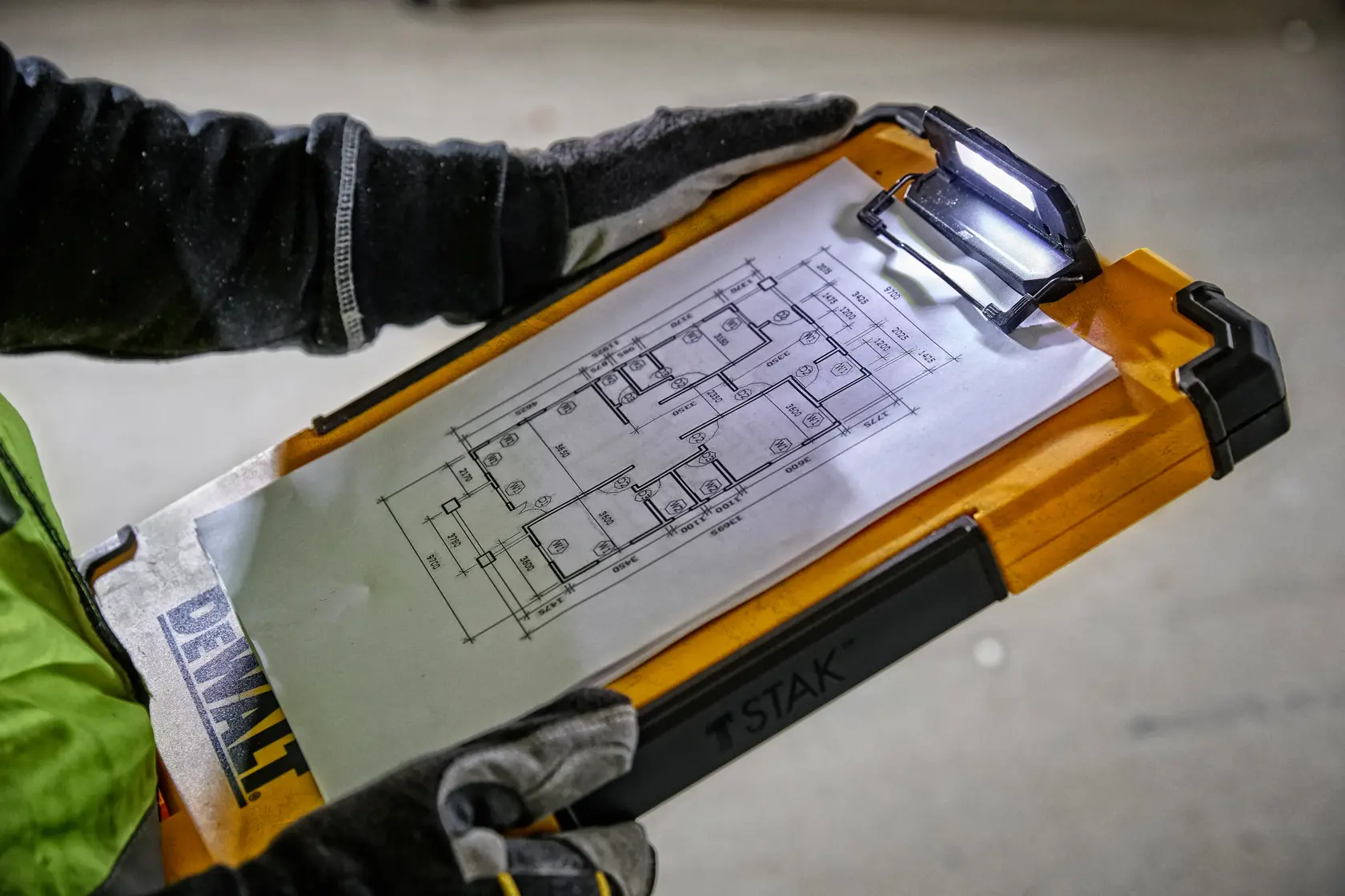 Eine Person mit Handschuhen hält ein DEWALT TSTAK Klemmbrett mit integrierter LED-Leuchte, auf dem ein technischer Grundriss an der Klammer befestigt ist.