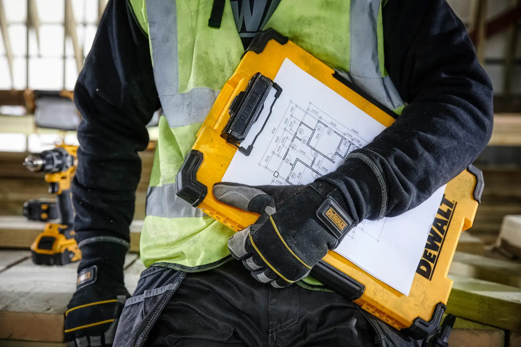 Eine Person mit Arbeitshandschuhen und Warnweste hält einen gelben DEWALT-Klemmbrettkoffer mit befestigten Bauzeichnungen auf einer Baustelle.