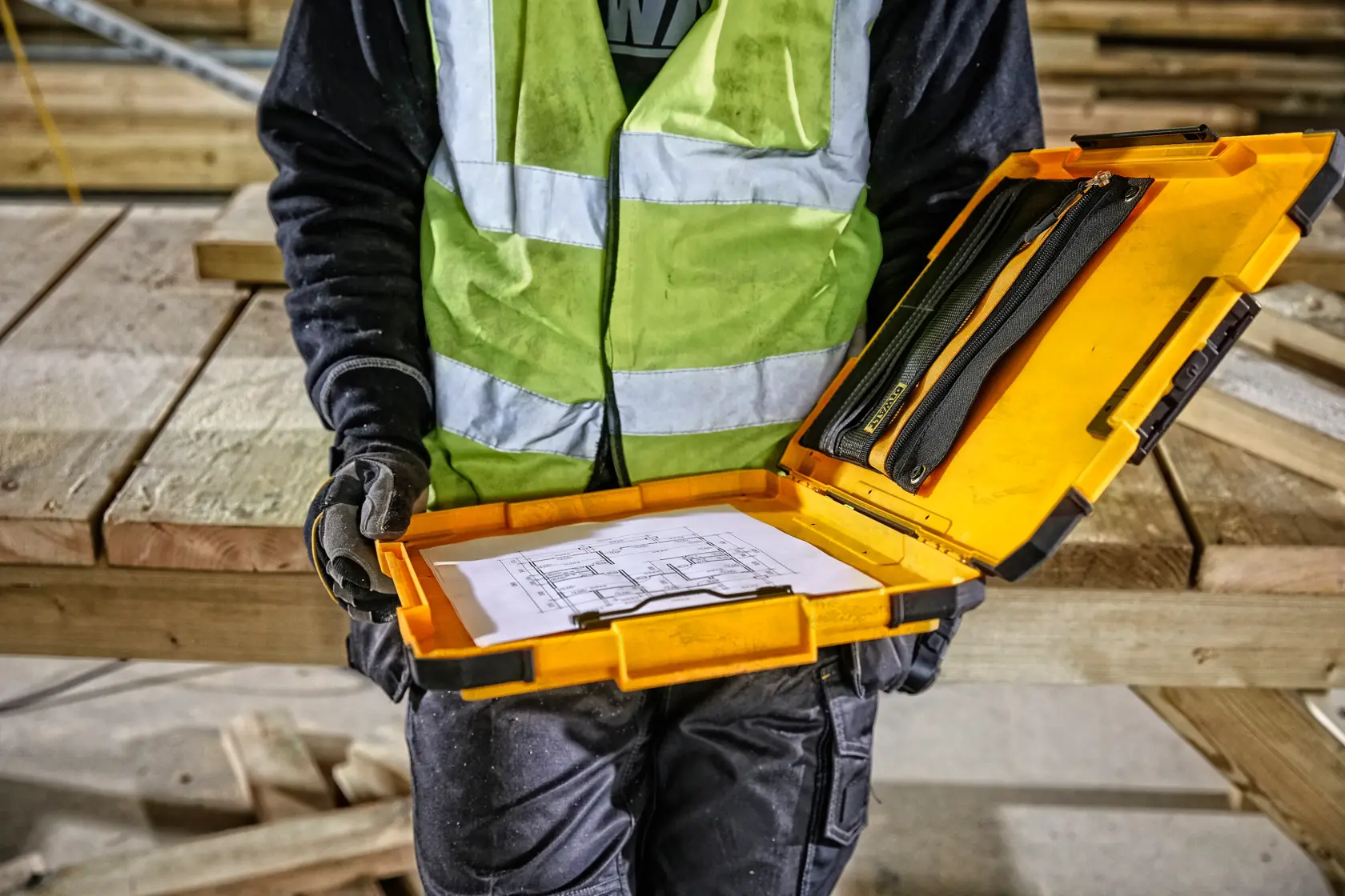 Eine Person mit einer reflektierenden Sicherheitsweste hält einen offenen gelben DEWALT-Koffer mit Dokumenten und Werkzeugen auf einer Baustelle mit Holzbalken im Hintergrund.
