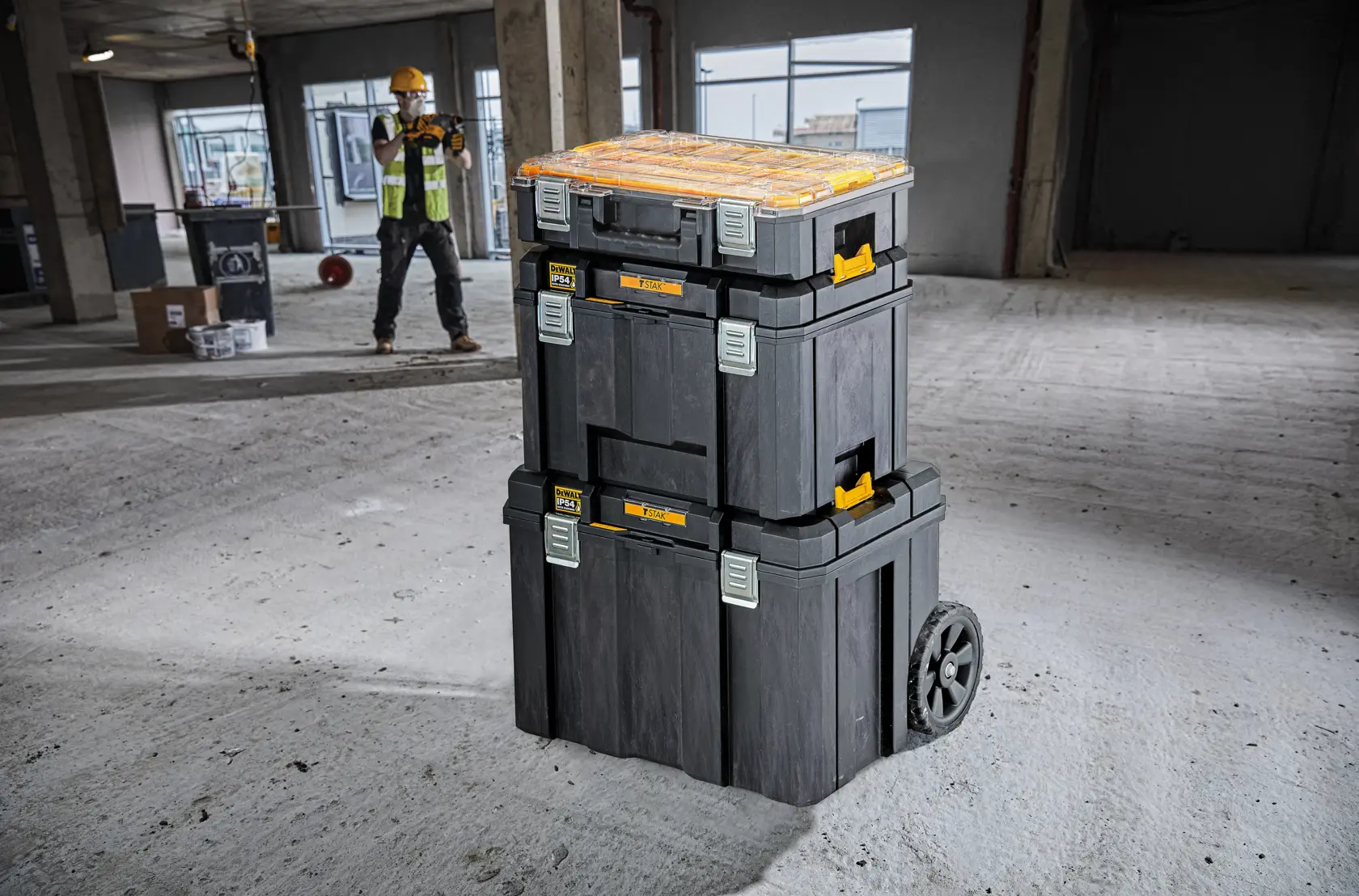 Cajas de herramientas DEWALT apiladas con ruedas en obra, trabajador de fondo.