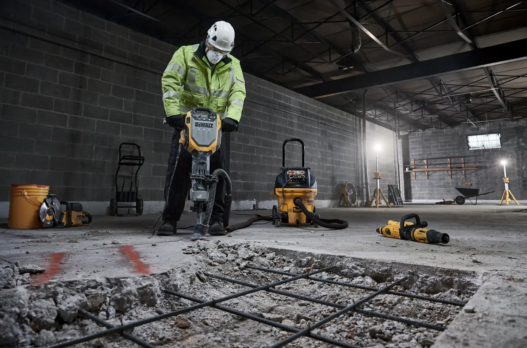 Arbeiter bricht Beton mit DEWALT Werkzeug, Staubabsauger und Ausrüstung stehen in der Nähe.