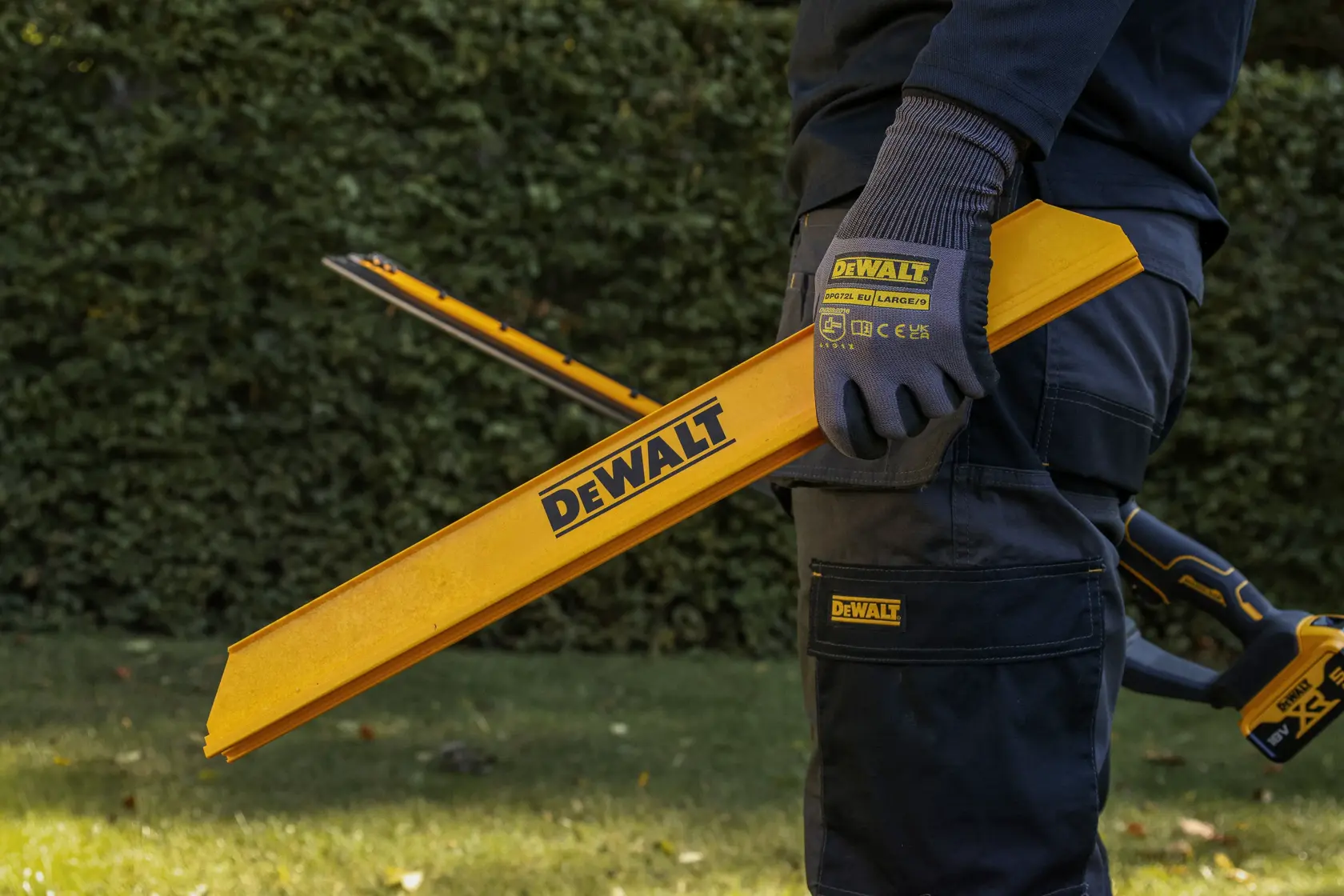 Person im Freien hält gelbes DEWALT-Werkzeug, trägt Handschuhe und Arbeitshose. Grüne Hecke im Hintergrund.